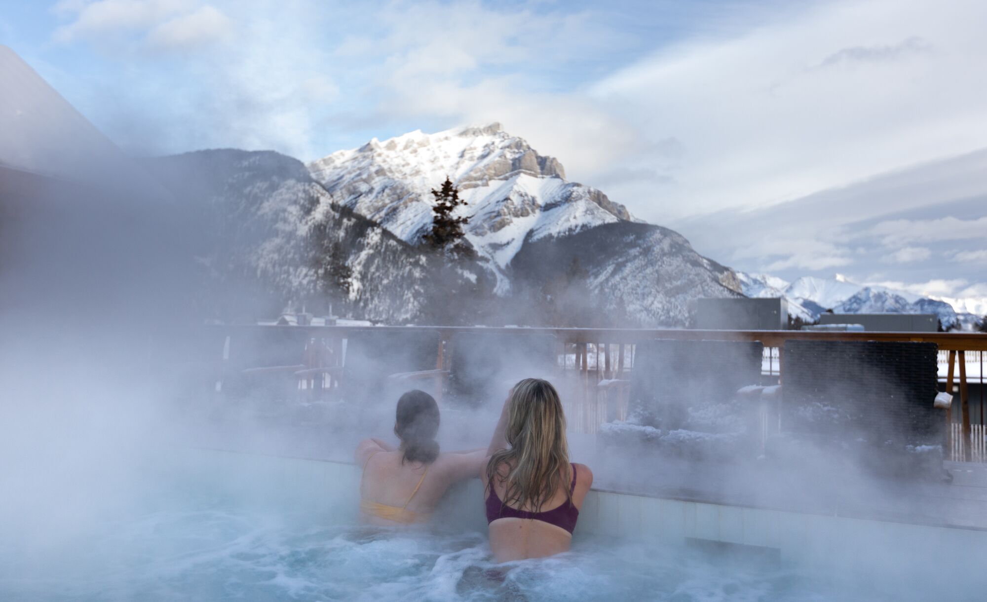 Two friends look at Cascade Mountain while enjoying a rooftop hot tub at the Moose Hotel in Banff.